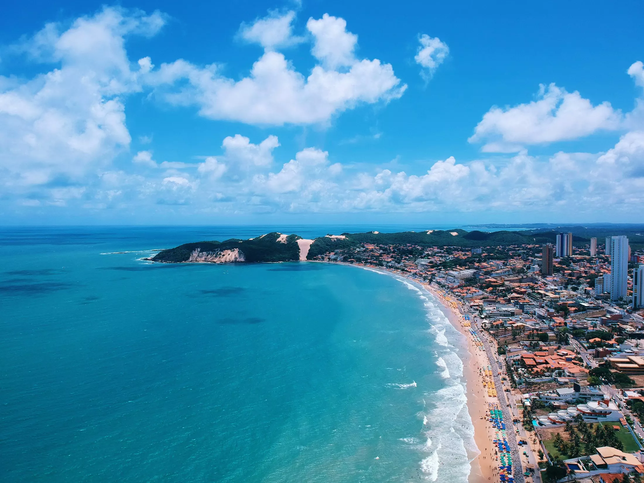 Vista aérea da orla de Natal, com prédios, a praia de areia clara e o mar azul